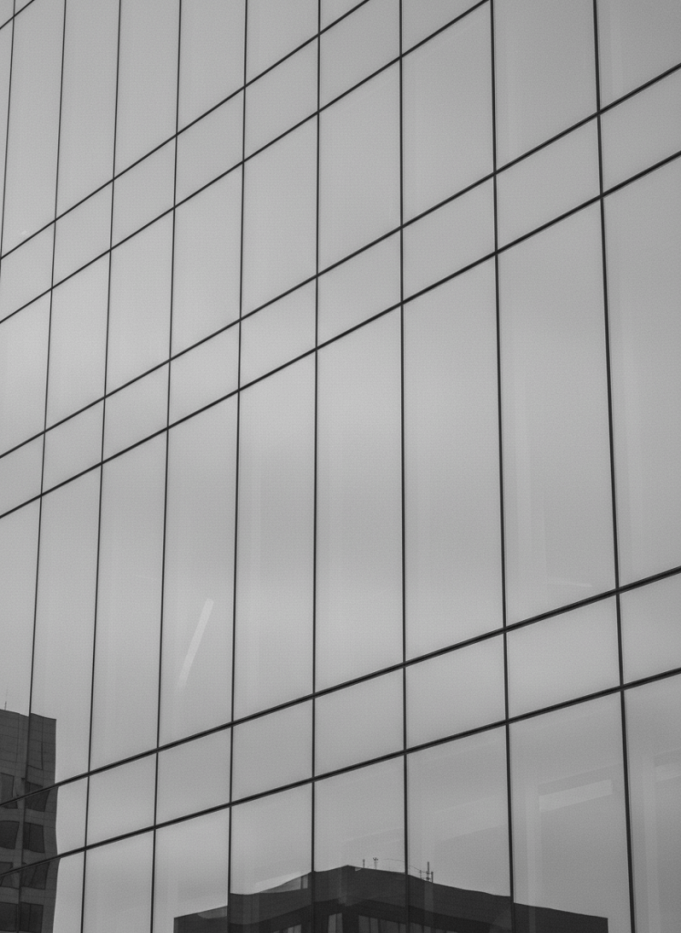 Muted light gray monochrome photograph of a modern glass office building facade with subtle reflections, soft low-contrast tones, clean minimal corporate aesthetic, faded washed-out grayscale